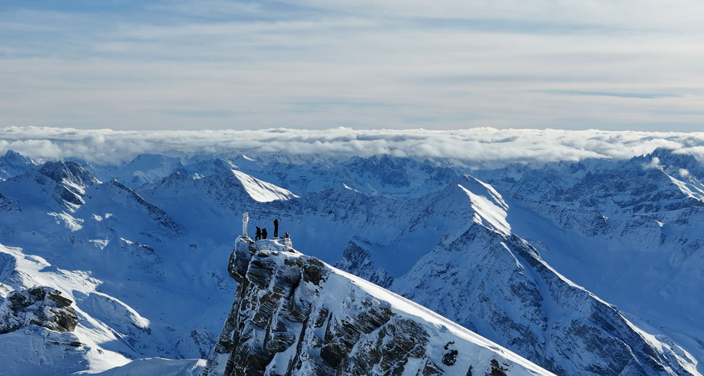 vue drone de 4 skieurs en haut du mont salsa, falaises en à-pics, croix sommital et panorama magnifique des alpes italienne dans l'arrière plan