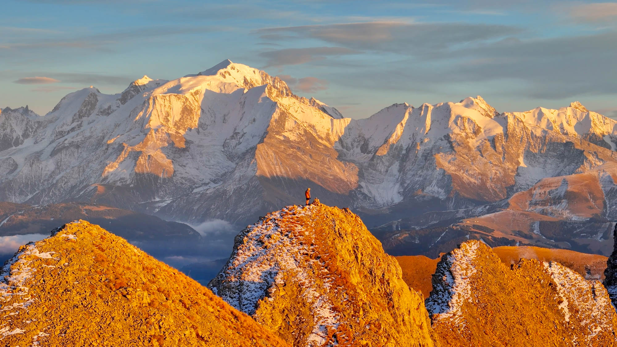 Silhouette au sommet d'une combe des Aravis dans une lueur orange de fin de journée, prise de vue au drone avec une vue sur le massif du mont blanc et le refuge du gouter
